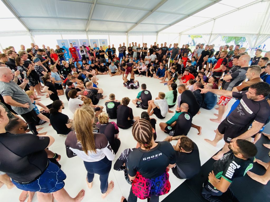 A large no-gi session in rash guards and shorts, seated in a circle around the instructor on the white competition mat