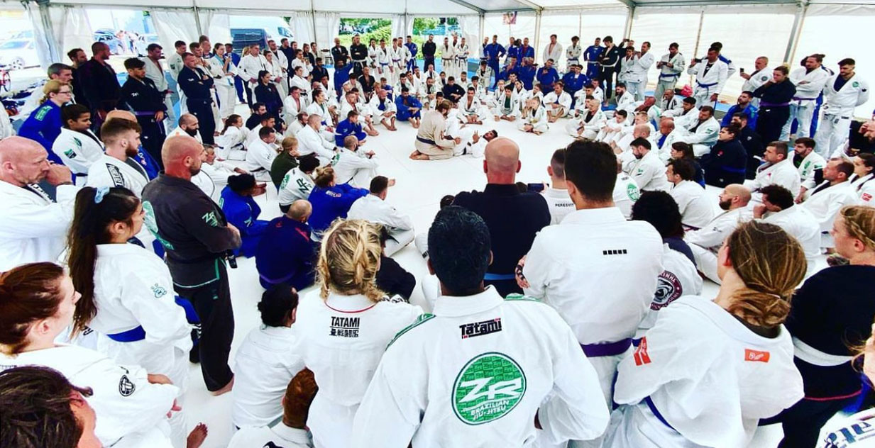 Camp participants in gi seated around an instructor teaching in the centre of the mat